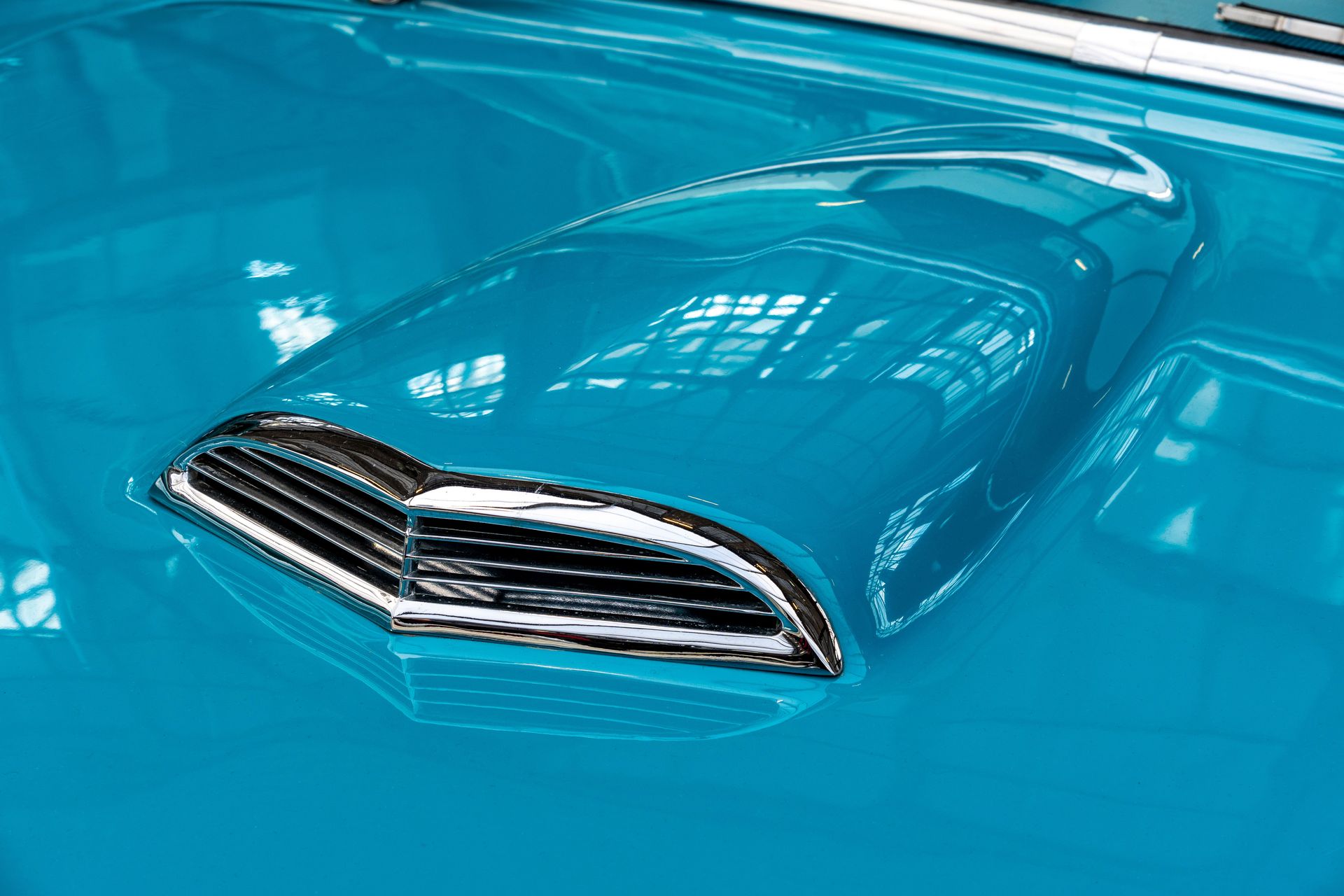 Detailed view of a blue vintage car hood featuring chrome accents, reflecting indoor lighting.